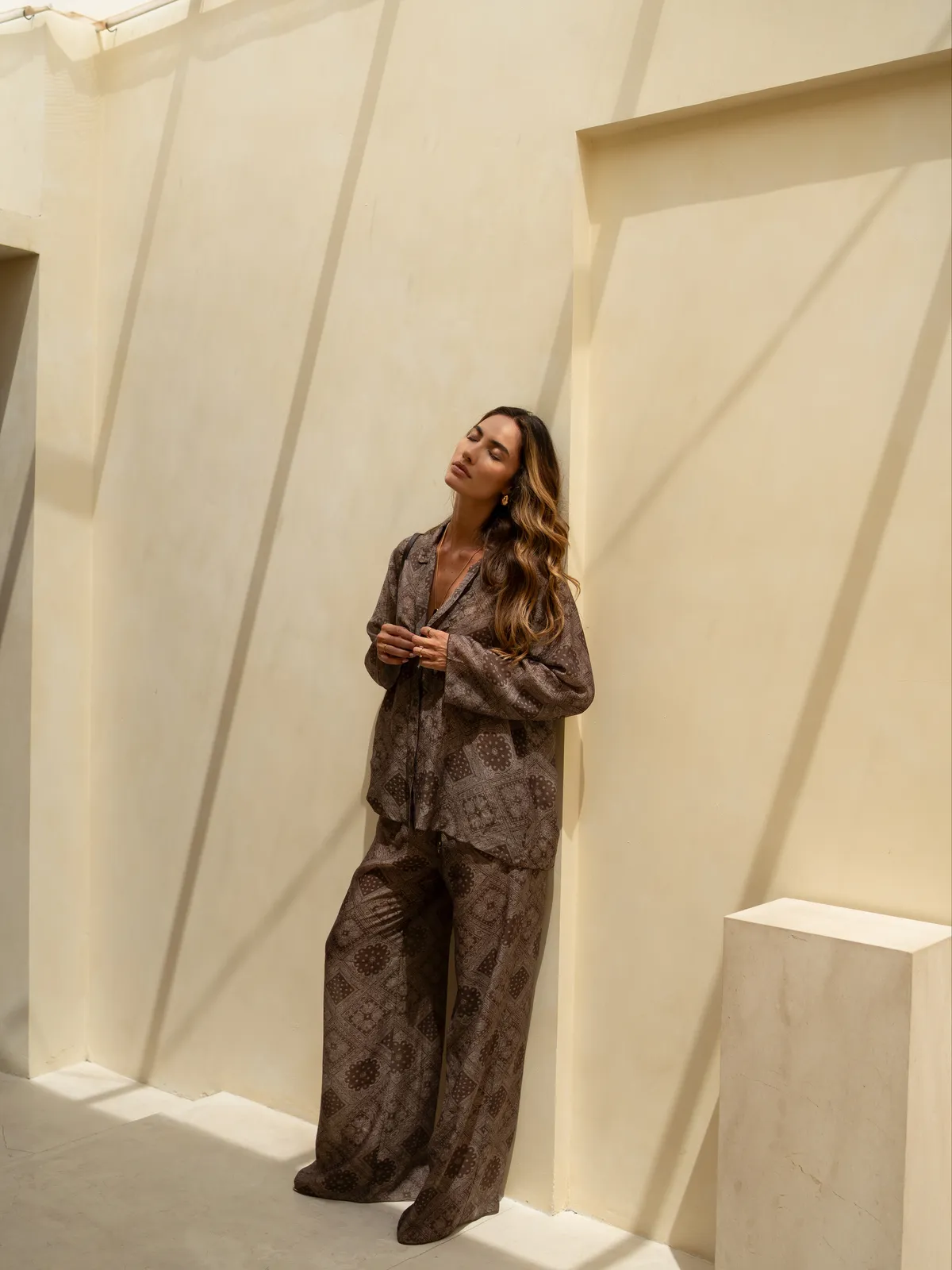 Модель у коричневому костюмі Suit Le Pyjama Brun MOODSTORIES з візерунком, що складається з вільної сорочки-жакета та широких штанів, стоїть біля дверей.