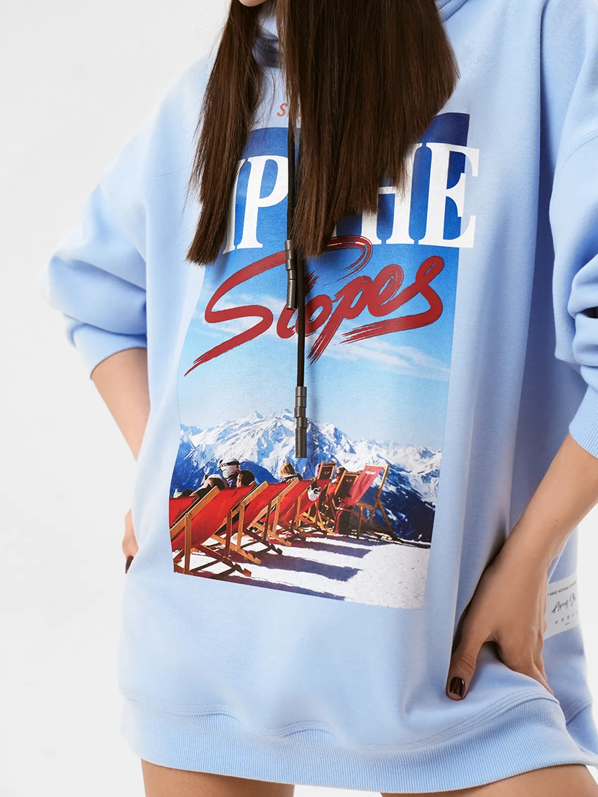 Модель в светло-голубом оверсайз свитшоте MOODSTORIES CALMA ALPINA с принтом "SIP THE Slopes" и белой пушистой шапке, держит сноуборд.