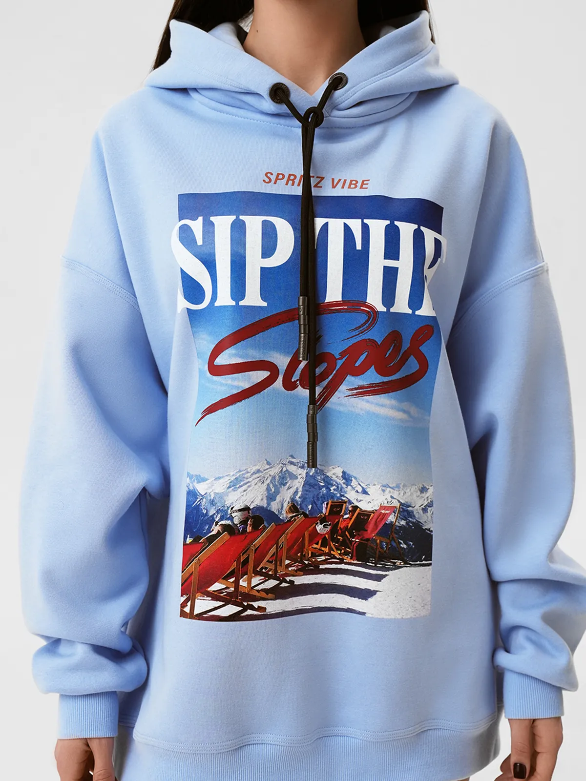 Модель в светло-голубом оверсайз свитшоте MOODSTORIES CALMA ALPINA с принтом "SIP THE Slopes" и белой пушистой шапке, держит сноуборд.