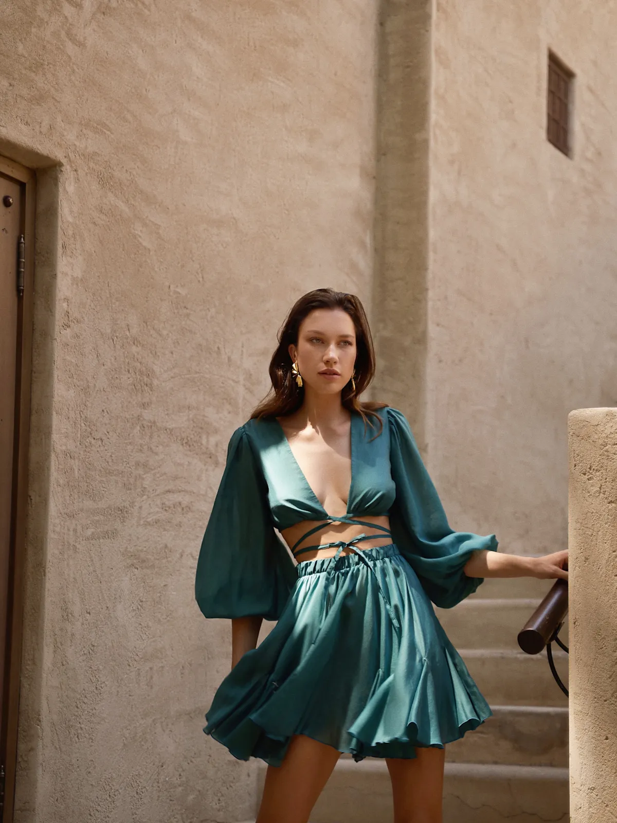 Модель в комплекте Suit Malibu Emerald от MOODSTORIES, состоящем из укороченного топа и мини-юбки, сидит на террасе.