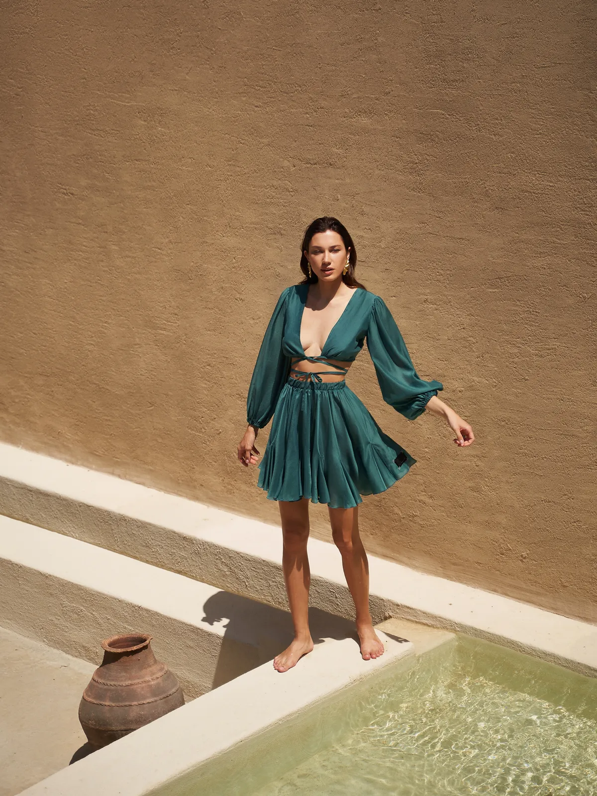Модель в комплекте Suit Malibu Emerald от MOODSTORIES, состоящем из укороченного топа и мини-юбки, сидит на террасе.